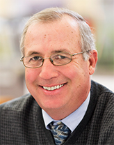 Joe Herr smiling, wearing a shirt, tie, and vest. 