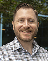 Kyle smiling in front of a garden in a plaid shirt.