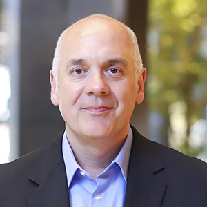 Joe wearing a navy blazer and blue shirt in front of a blurred background.