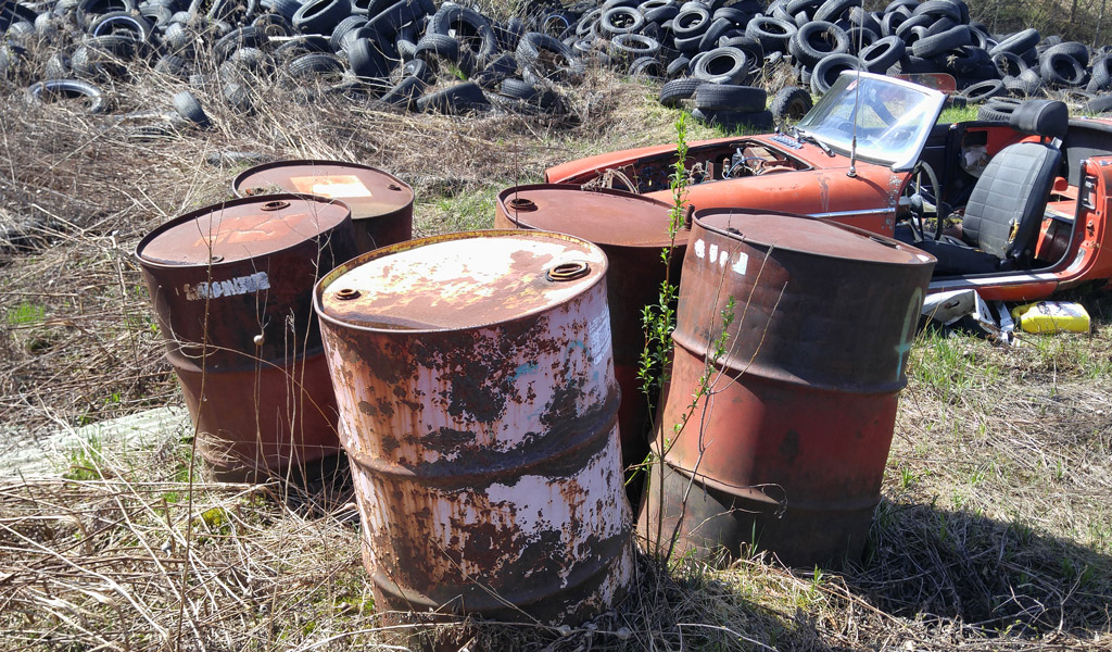 Grassy lot with rusted barrels, scrapped car, and large pile of tires.