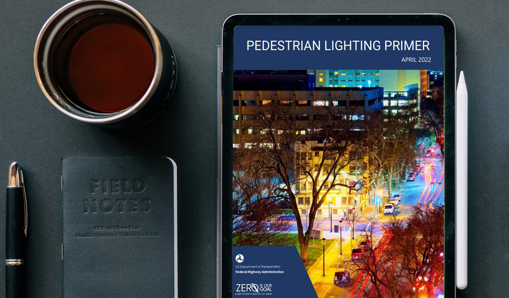 Next to a pen, coffee cup, and notebook, an iPad on a desk features the Pedestrian Lighting Primer screen.