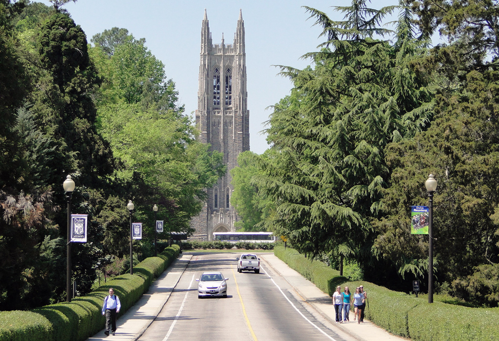 The road leading into Duke University, with people walking on sidewalks and the campus in the background.