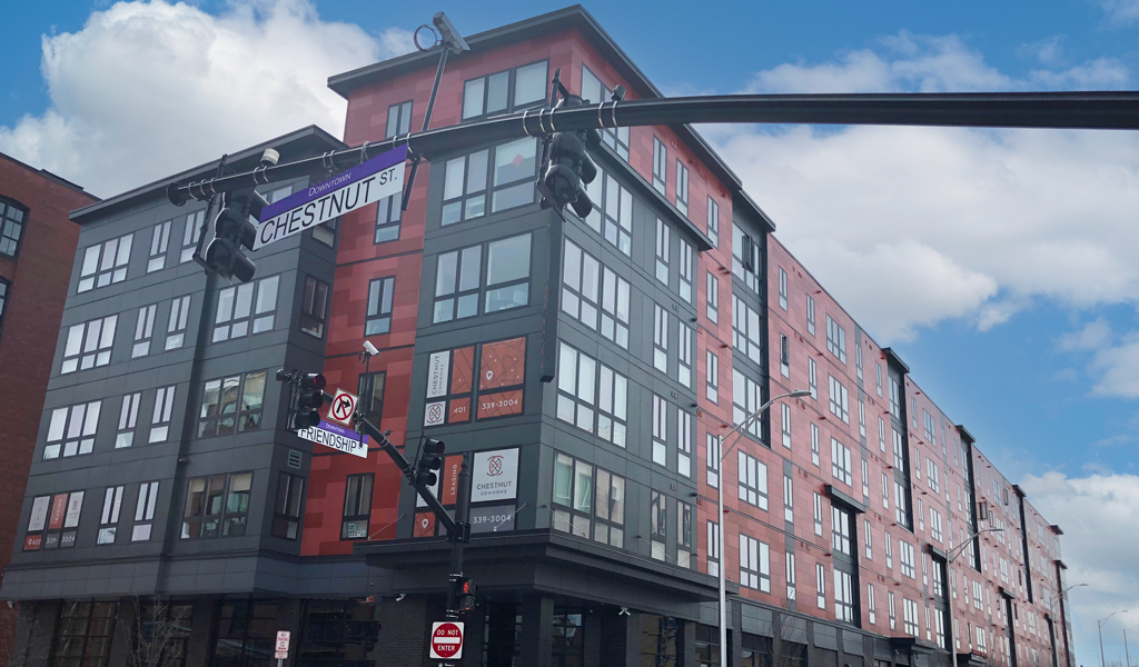 Urban street view of a modern six-story building featuring red and gray exterior and parked cars along sidewalk.