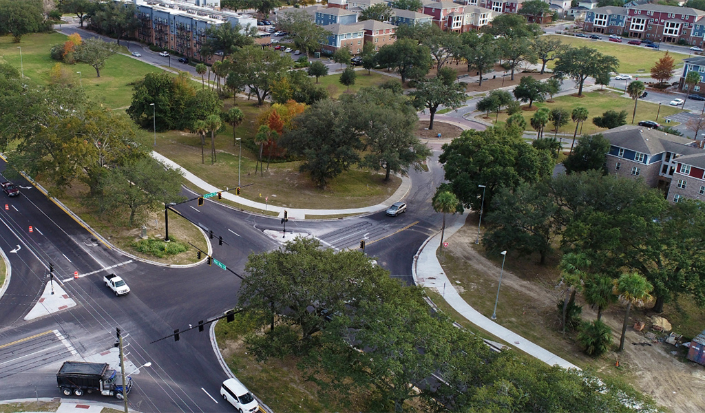 Cars drive past a tree-shaded raised median seen from above. 