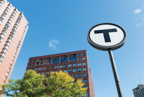 Cityscape with a prominent Boston MBTA transit system 'T' sign, surrounded by modern buildings and a clear blue sky.