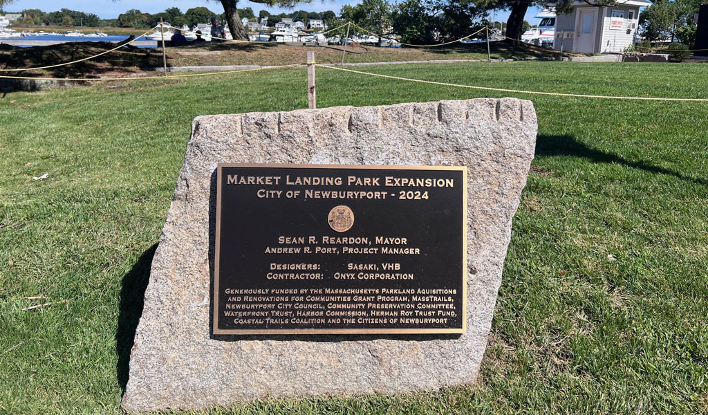 Commemorative plaque for the Market Landing Park Expansion displayed on a stone in a grassy area with trees and a blue sky.