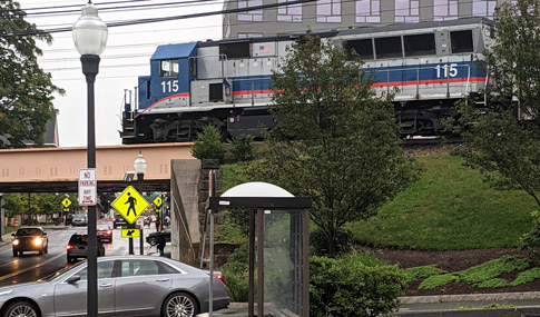 A train car on tracks above a street that is busy with cars. A bus stop is on the sidewalk.