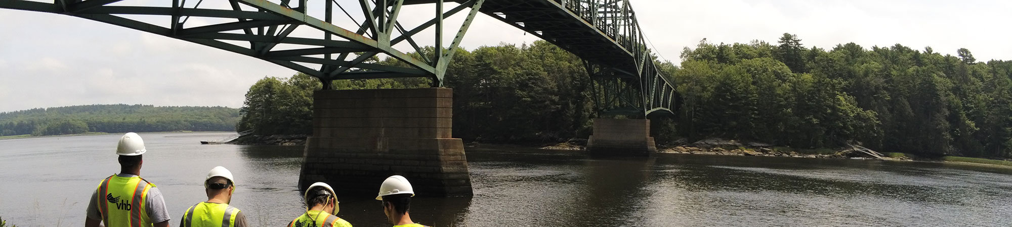 A group of men use an unmanned drone under a bridge crossing over a large river.