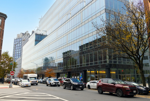 Modern glass lab building in New York with parked cars and traffic along a busy city street on a clear fall day.