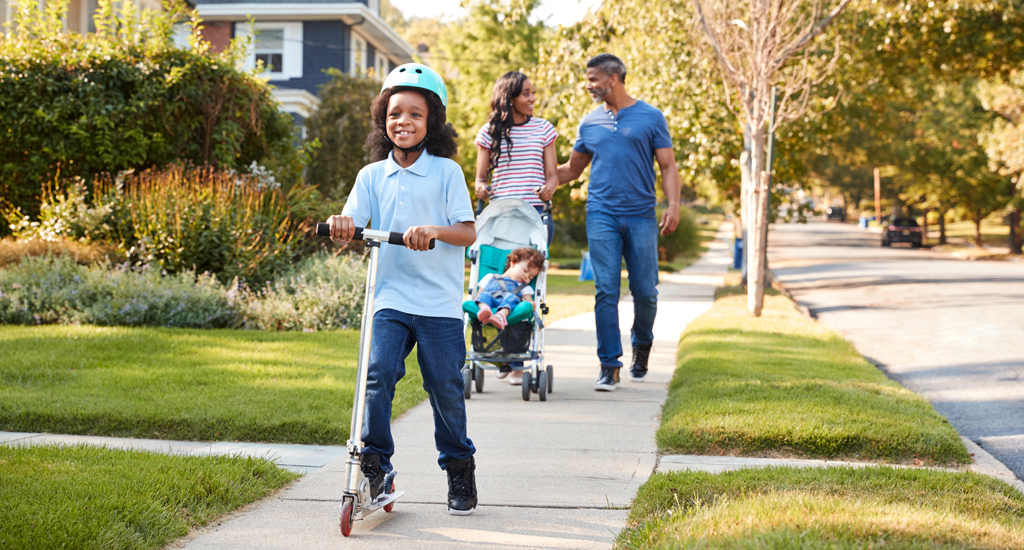 A child on a scooter with their family walking behind them on a sidewalk.