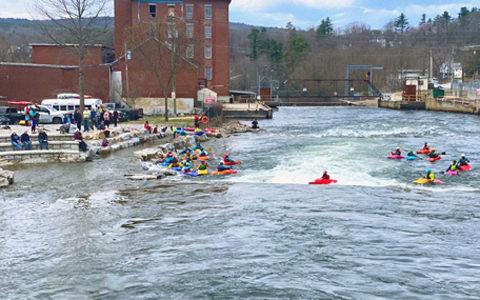 Group of kayakers paddling through rapids in a river, with spectators watching from the riverbank and surrounding areas in a small-town setting.