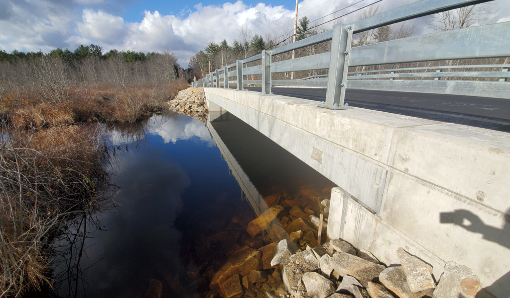 Concrete bridge over a clear stream with visible rocks underneath, surrounded by dried vegetation under a partly cloudy sky.