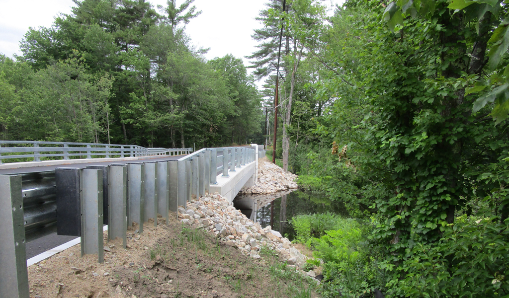 View of a newly constructed bridge surrounded by lush greenery, with a clear sky above and a stream flowing underneath. 
