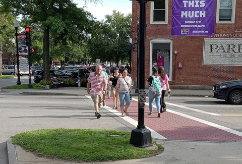 Pedestrians crossing a street in downtown Keene, NH near a signpost and Parrish Shoes wall mural.