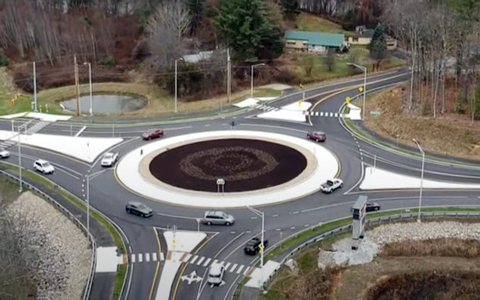 Aerial view of a traffic roundabout with multiple exits and vehicles. The roundabout features a decorative center with landscaping.