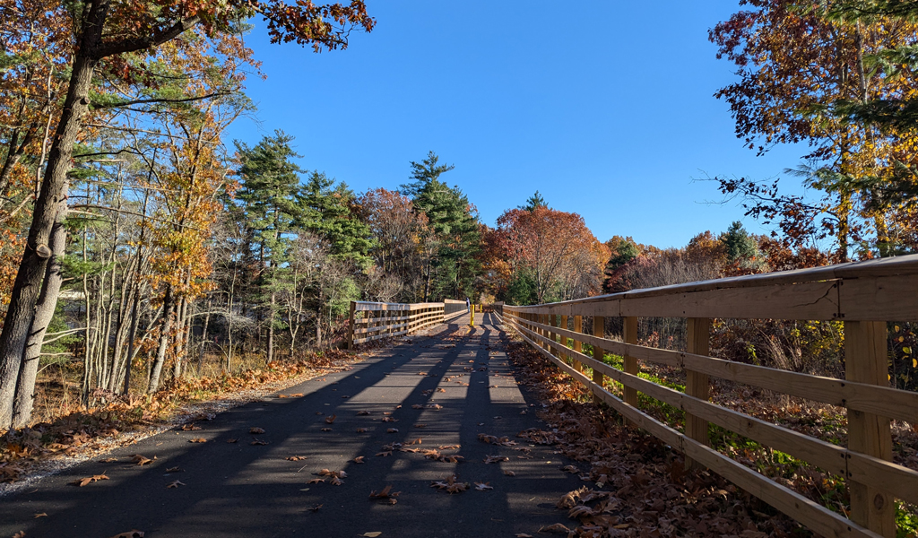 Autumn scenery along a paved path with a wooden fence on one side, surrounded by trees with fall foliage under a blue sky.