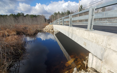 View of a concrete bridge over a tranquil river with clear reflections under a partly cloudy sky.