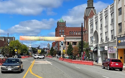View of Main Street in Nashua, NH with a banner reading "Welcome" across the road, flanked by historic buildings under a partly cloudy sky.