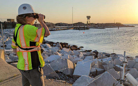 Anna Romano looks through binoculars as she bird monitors during a site visit.