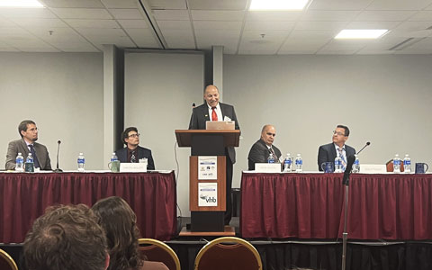 A man wearing a business suit speaking at a podium with the NHF and VHB logos on it, alongside four seated panelists at a conference event in a room with banners displaying the NHF logo.