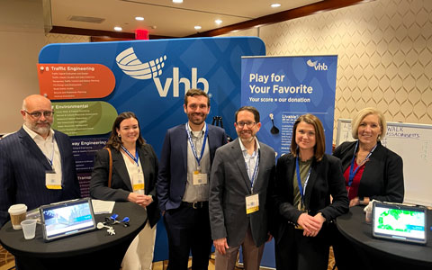 Jim Kersten, Katie Mancinelli, Matt Duranleau, Lourenço Dantas, Niki Hastings, and Carrie McInerney stand together smiling in front of the VHB booth at the Moving Together Conference wearing business attire. 