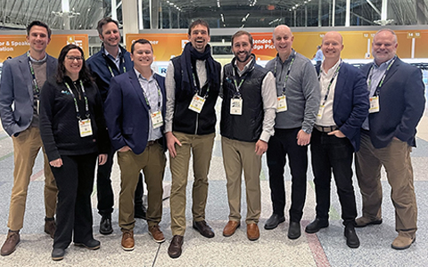 Group of ten professionals smiling for a photo at a conference with banners in the background, including a logo for "Partner & International Education". They are wearing name badges and business casual attire.