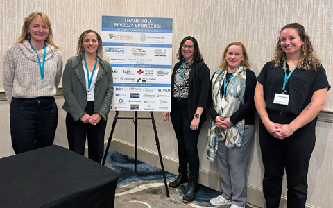 VHBer stand together wearing business attire in front of a sign thanking the 2024 REV Conference sponsors. 