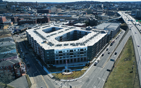 Aerial view of a large, modern apartment building with a U-shaped design, located near a busy highway and surrounded by various other buildings under clear skies.