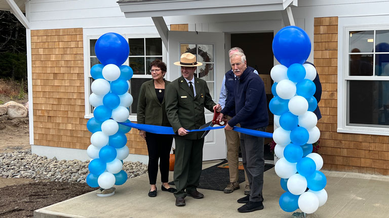 Three people participating in a ribbon-cutting ceremony in front of a new building, decorated with blue and white balloons. A camera is positioned in the foreground, capturing the event.