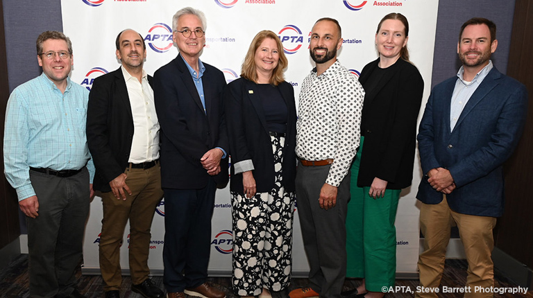 Group of seven professionals standing together at an APTA (American Public Transportation Association) event, smiling for the camera in front of a backdrop with APTA logos.