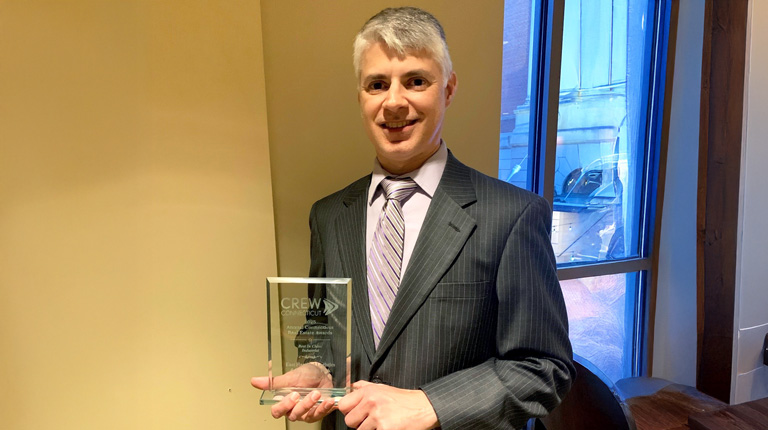 Person holding a CREW Network award, wearing a suit and tie, smiling in an indoor setting.