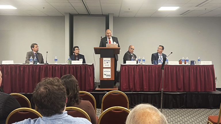 A man wearing a business suit speaking at a podium with the NHF and VHB logos on it, alongside four seated panelists at a conference event in a room with banners displaying the NHF logo.