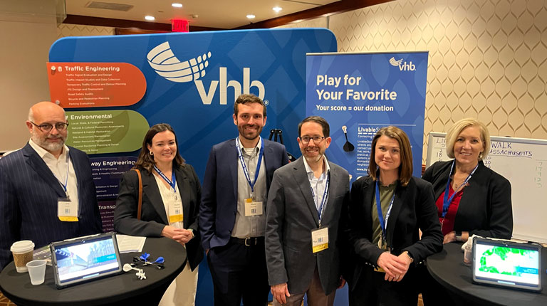 Jim Kersten, Katie Mancinelli, Matt Duranleau, Lourenço Dantas, Niki Hastings, and Carrie McInerney stand together smiling in front of the VHB booth at the Moving Together Conference wearing business attire. 