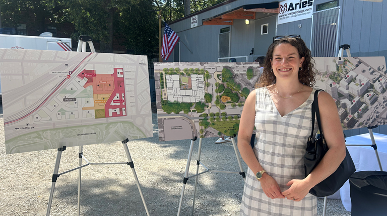 A person standing in front of three informational display boards showing urban development plans, with a building and American flags in the background.
