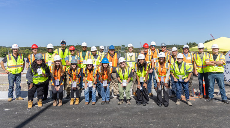 Group of construction workers and students in safety gear standing together in a line at a construction site. 