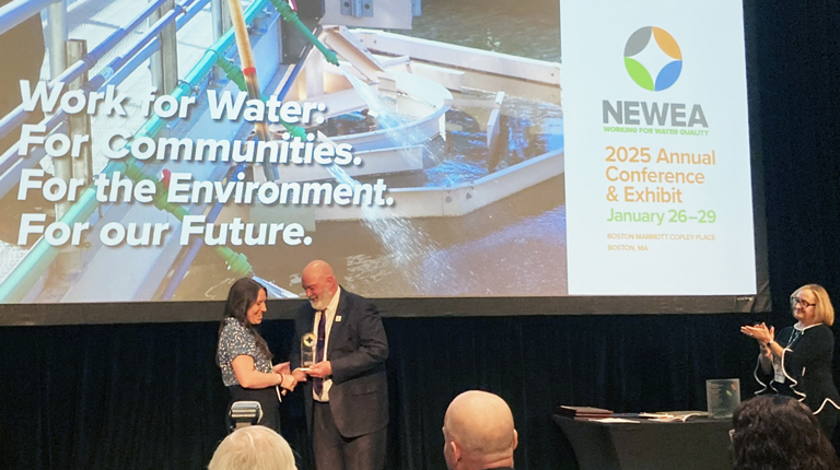 Two individuals shaking hands on stage at the NEWWA 2025 Annual Conference & Exhibit, with a backdrop displaying the slogan 'Work for Water. For Communities. For the Environment. For Our Future.' and images of water treatment infrastructure.