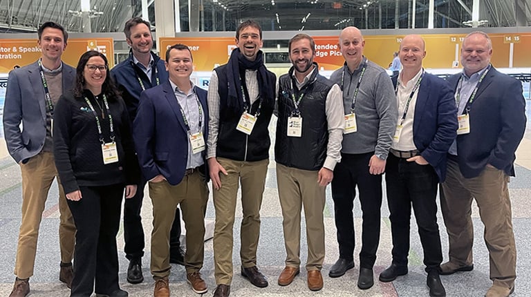 Group of ten professionals smiling for a photo at a conference with banners in the background, including a logo for "Partner & International Education". They are wearing name badges and business casual attire.