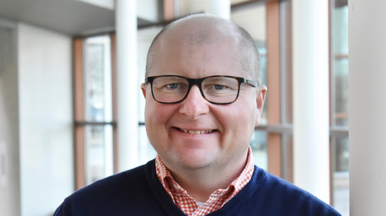 Headshot of Kurt Muller wearing business casual attire smiling in the lobby of an office building.