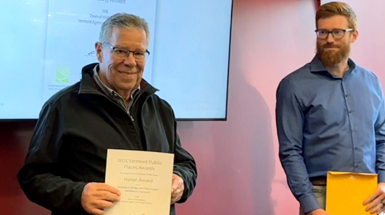 Two individuals are smiling and standing in front of a presentation screen displaying the "2025 Vermont Public Places Awards." 