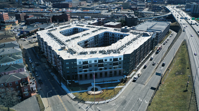 Aerial view of a large, modern apartment building with a U-shaped design, located near a busy highway and surrounded by various other buildings under clear skies.