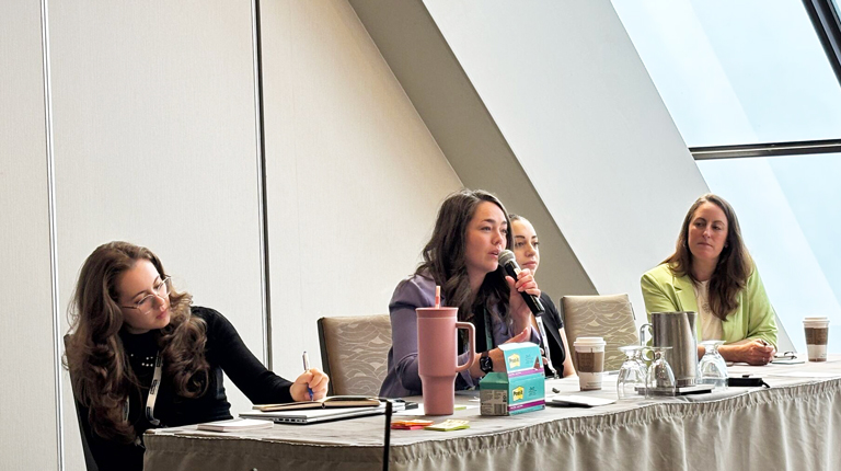 Four individuals participate in a panel discussion at a conference room table, equipped with microphones and laptops.