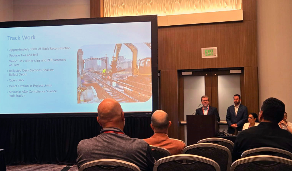 Two presenters at a conference discussing track reconstruction, with a slide presentation in the background showing construction images and bullet points outlining the project details.