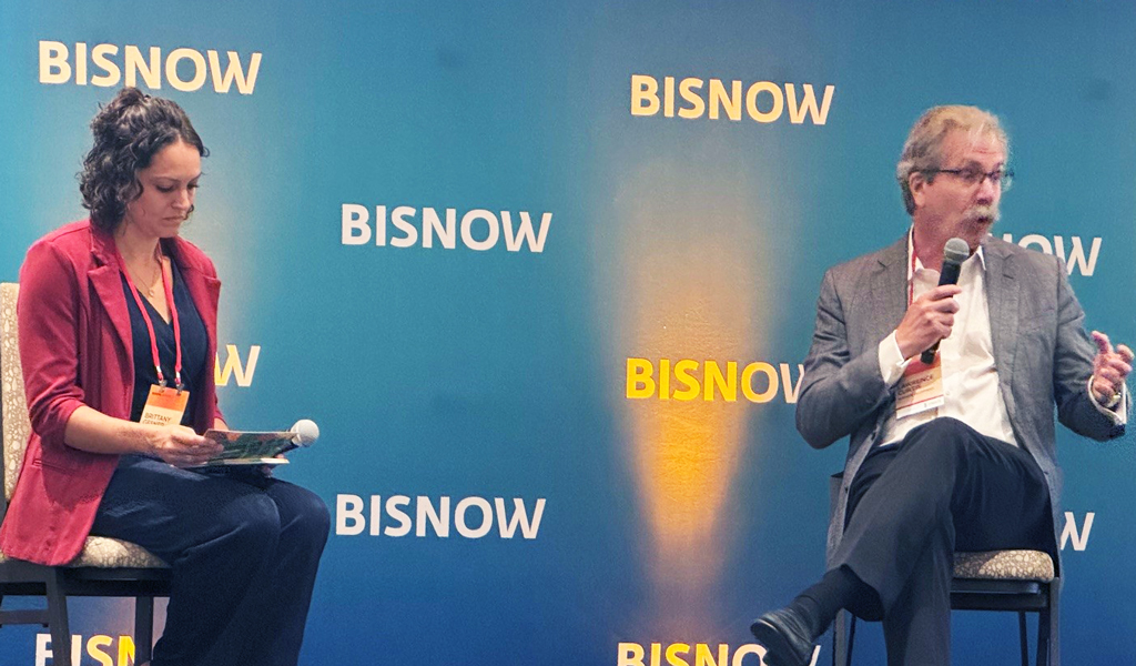 Two individuals participating in a panel discussion at a Bisnow event. One is seated on the left, looking at notes, and the other is holding a microphone, speaking animatedly.