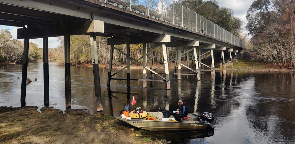 Chris Crow is in a boat under a bridge conducing a mussel survey.