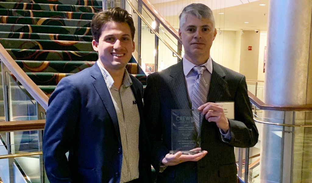 Two people at a formal event with a staircase in the background. One person is holding the CREW Network award.