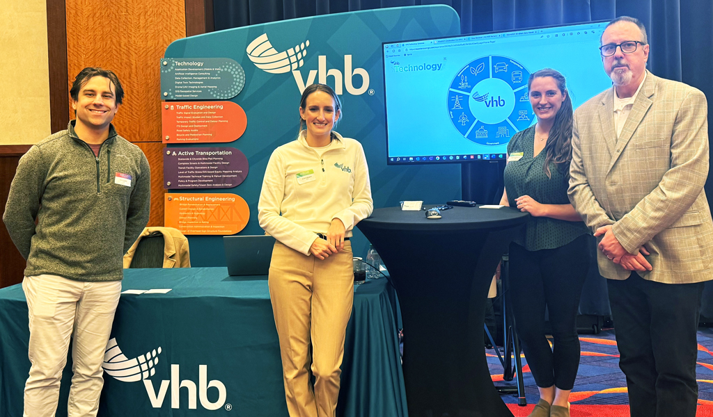 Four VHB employees standing in front of an exhibit booth featuring the VHB logo.