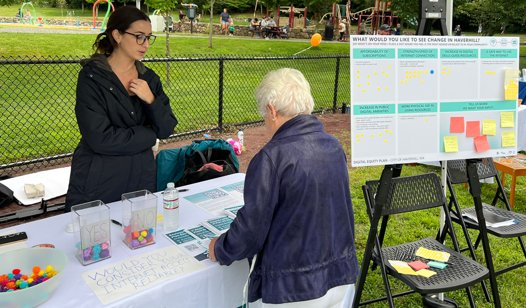 A VHB popup tent and table at the Farmers Market with a woman in a black coat speaking to a gentleman in a gray sweatshirt. 