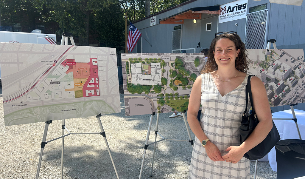 A person standing in front of three informational display boards showing urban development plans, with a building and American flags in the background.