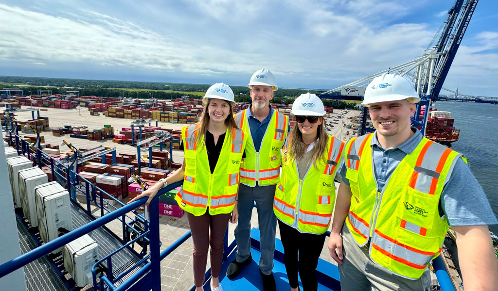 People wearing hard hats and standing on a crane above a port.