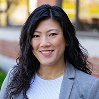 A person with long curly hair, wearing a light gray blazer and a white shirt, is positioned against a black background.